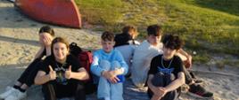 Czterech chłopców, dwie dziewczyny siedzą na piasku nad brzegiem jeziora, za nimi odwrócona łódka