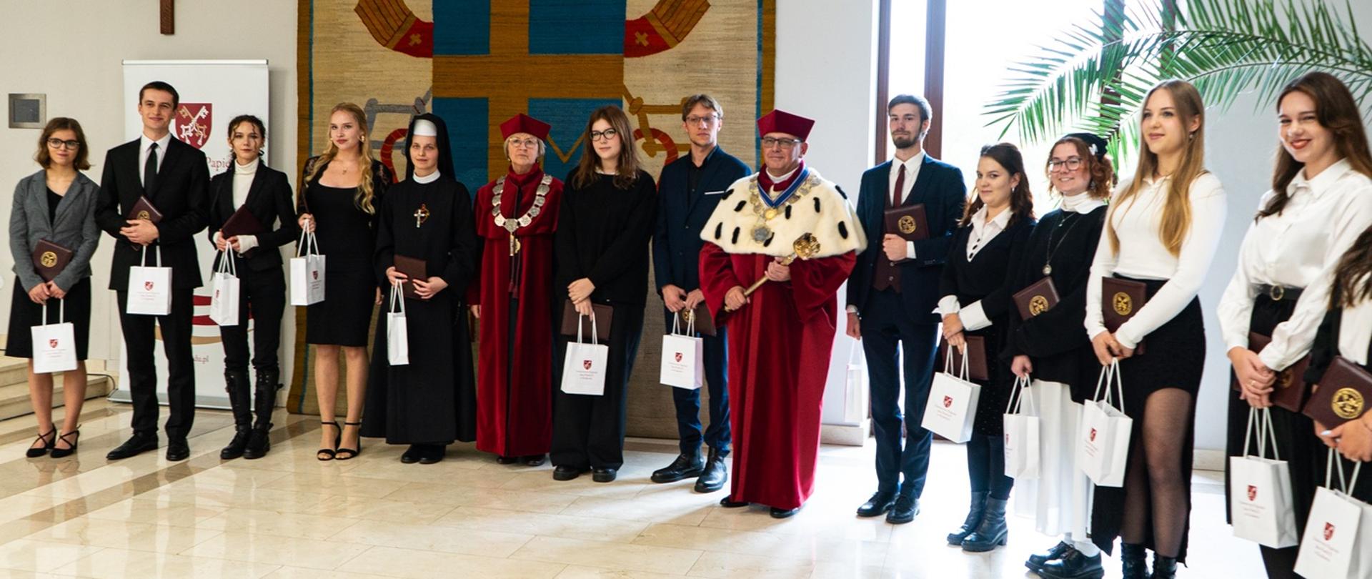 Do fotografii grupowej pozują studenci i profesorowie; na ścianie za nimi drewniany krzyż oraz duży herb papieski.