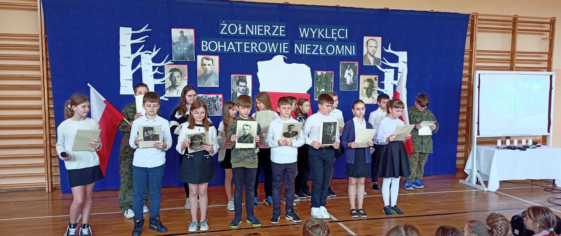 Uczniowie klasy piątej występują w akademii, trzymają w rękach zdjęcia przedstawiające żołnierzy wyklętych, ubrani są na galowo, stoją na tle granatowej dekoracji z mapą Polski i zdjęciami żołnierzy wyklętych