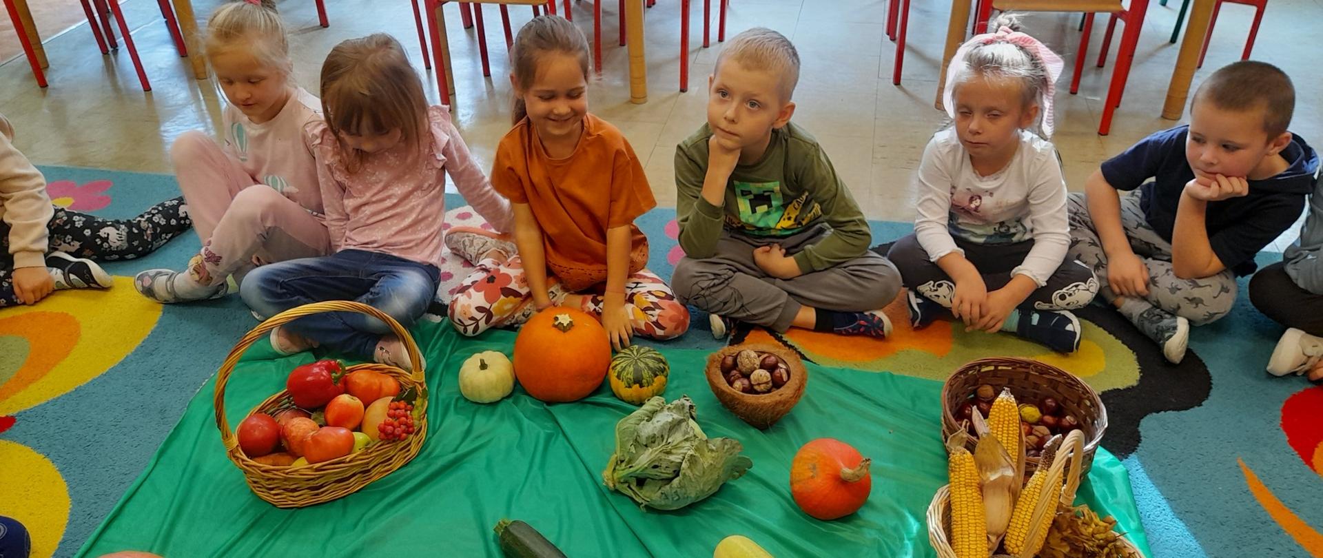 Dzieci siedzą na dywanie, przed nimi kosze z warzywami i owocami jesiennymi