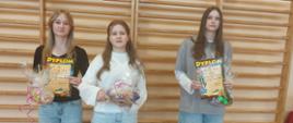 Trzy dziewczynki reprezentujące swoje drużyny stoją na sali gimnastycznej i trzymają w ręku nagrody i dyplomy