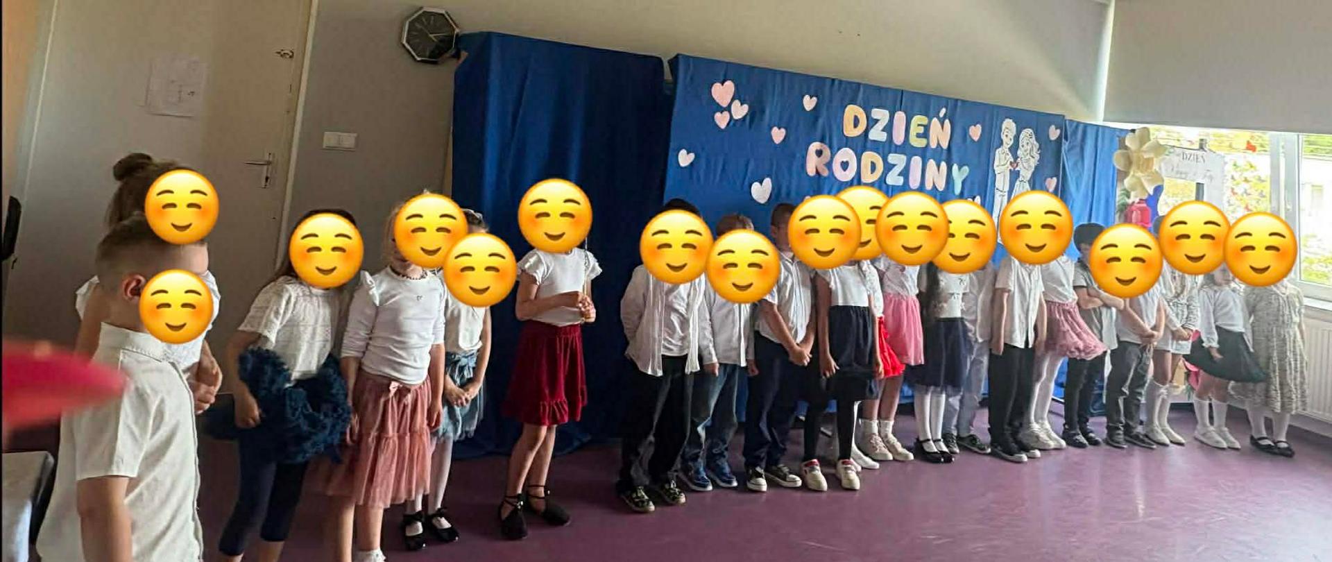 Dzieci ustawione w jednej linii. Wszystkie dzieci są elegancko ubrane i mają twarze zasłonięte emotikonami. Niebieskie tło z białymi napisami.