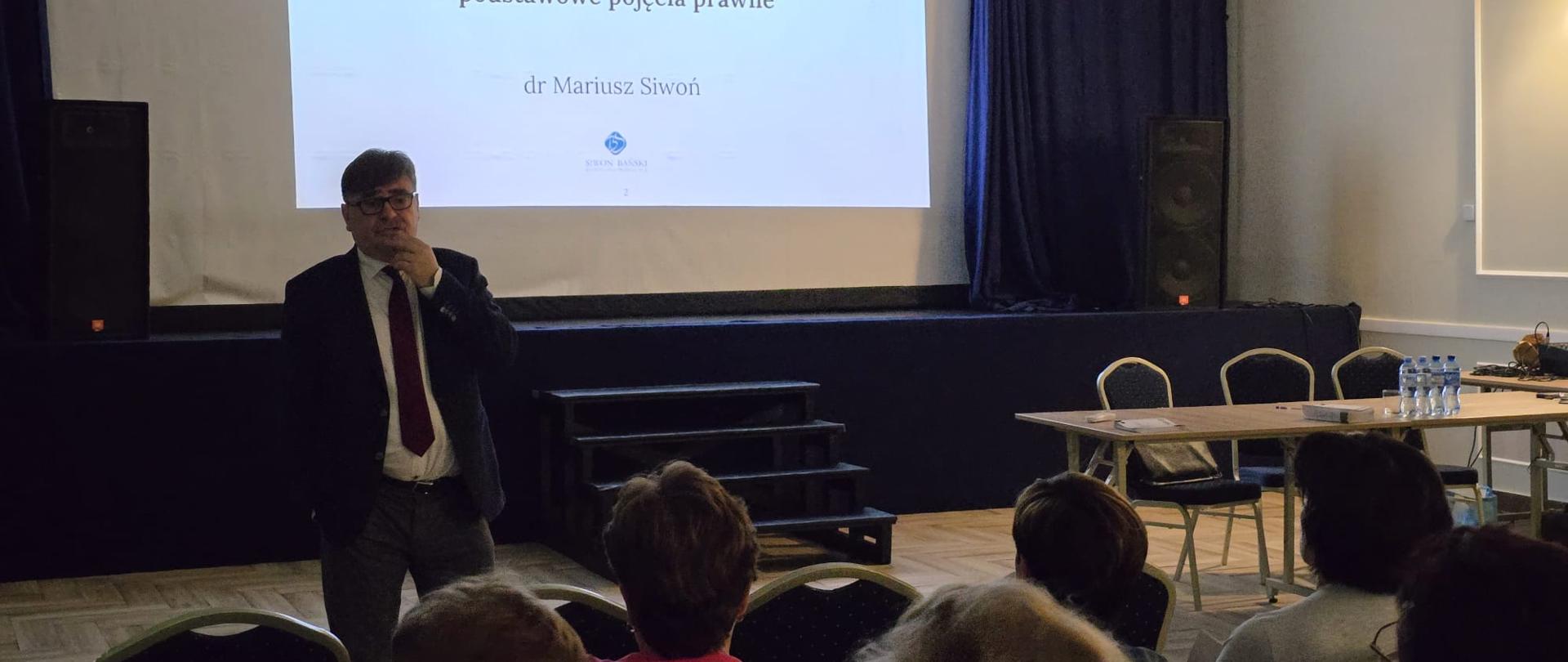 Mężczyzna stoi przed ekranem projekcyjnym słuchają go siedzący naprzeciw ludzie