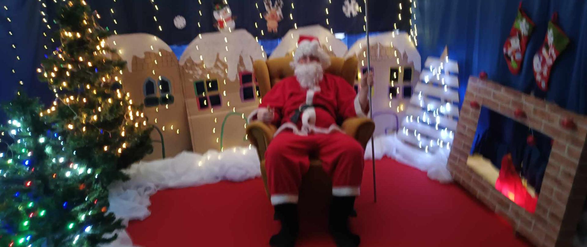 Święty Mikołaj siedzący w fotelu na tle dekoracji zimowej i oświetlonych choinek 