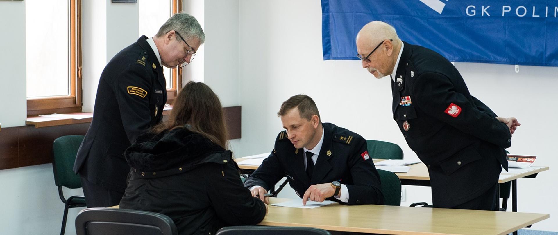 Na zdjęciu trzech strażaków wchodzących w skład komisji konkursowej zadaje pytania uczestniczce siedzącej w ławce na sali konferencyjnej w siedzibie Państwowej Straży Pożarnej w Sokołowie Podlaskim