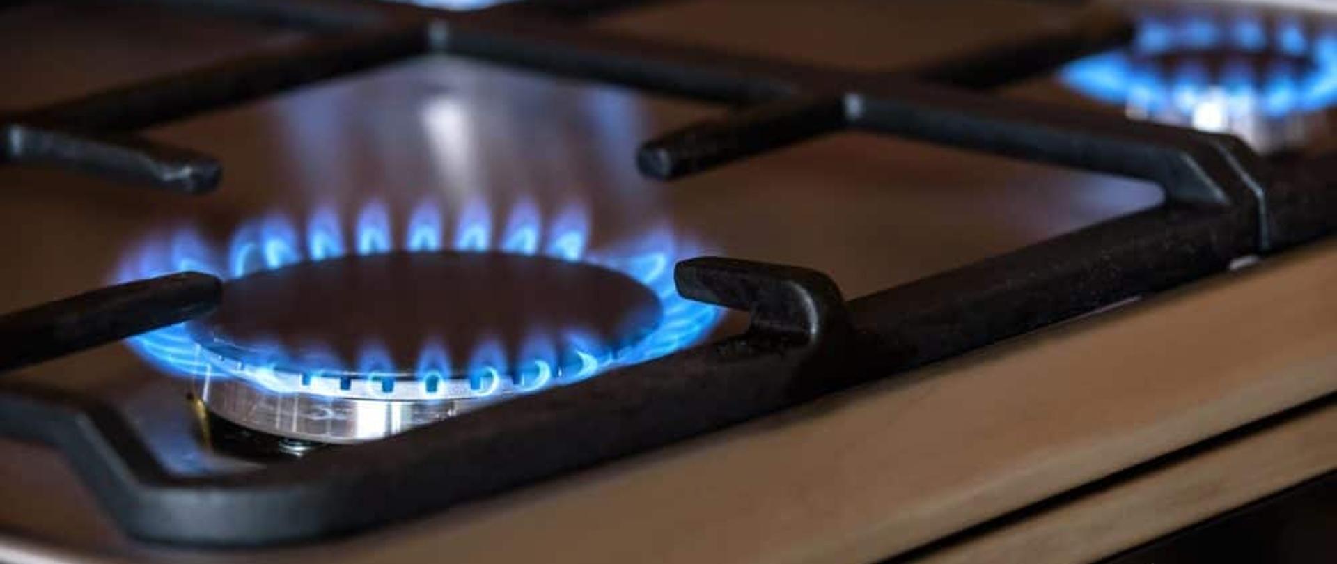 Na fotografii szara kuchenka gazowa na której odkręcony jest gaz. Na pierwszym planie niebieskie płomienie gazu. 