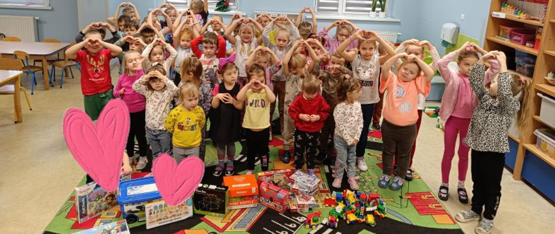 Grupa dzieci przedszkolnych z podniesionymi rączkami, dłonie ułożone w kształcie serduszka, przed nimi na dywanie leżą zabawki, samochodziki, puzzle i klocki. W tle trzy okna zasłonięte roletami. 