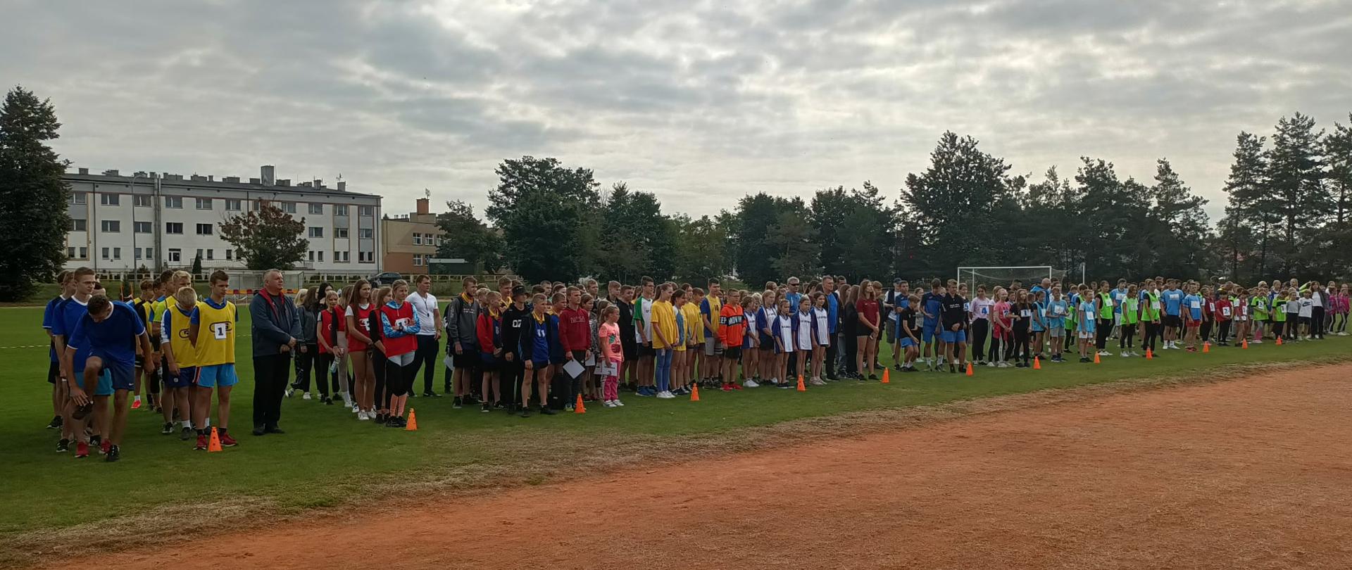 Inauguracja Szkolnego Roku Sportowego 2021/2022 na Stadionie Miejskim w Chorzelach. Fotografia przedstawia uczniów biorących udział w zawodach. Młodzież stoi na murawie boiska podzielona na grupy według płci, wieku oraz szkoły.
