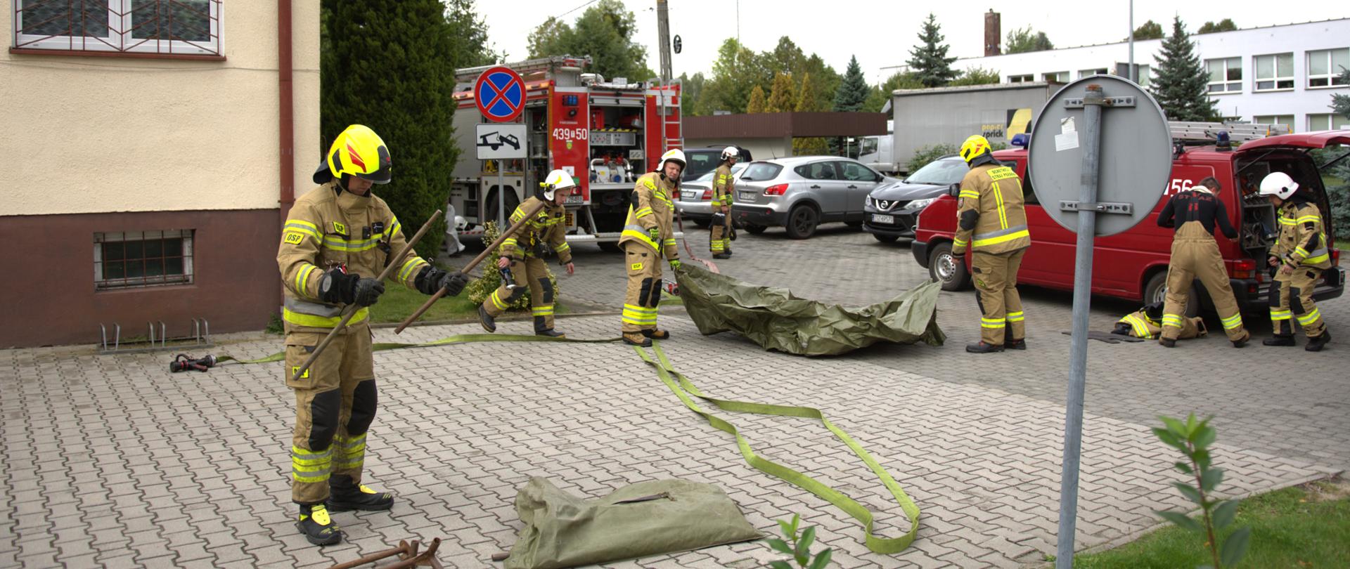 Na zdjęciu obok Urzędu Gminy w Rudzie Malenieckiej widać 7 strażaków. Znajdują się tam również 2 samochody strażackie. Strażacy rozkładają sprzęt strażacki i przygotowują się do realizacji akcji.