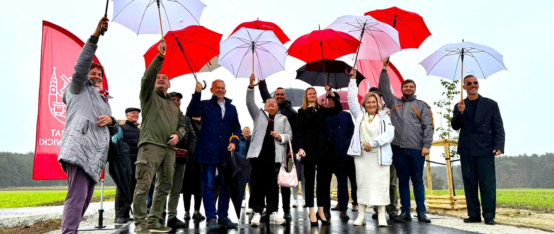 Grupa ludzi unosi nad głowami parasolki. Parasolki są w kolorach białym i czerwonym. W tle widać nowo otwartą drogę w miejscowości Brunów, gmina Chocianów