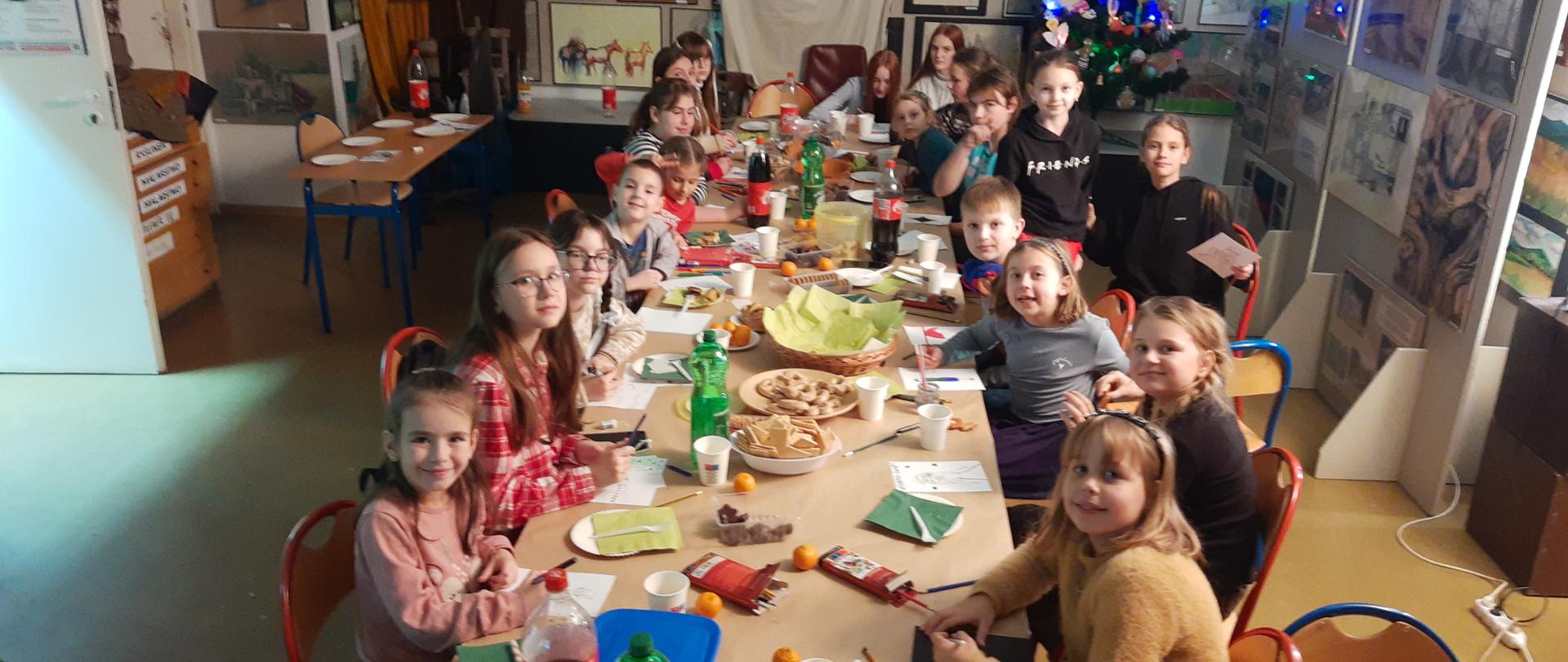 Na zdjęciu dzieci z Polski i Ukrainy siedzą przy wspólnym stole, w sali zajęć z rysunku i malarstwa, w tle stoi choinka ozdobiona pracami dzieci.