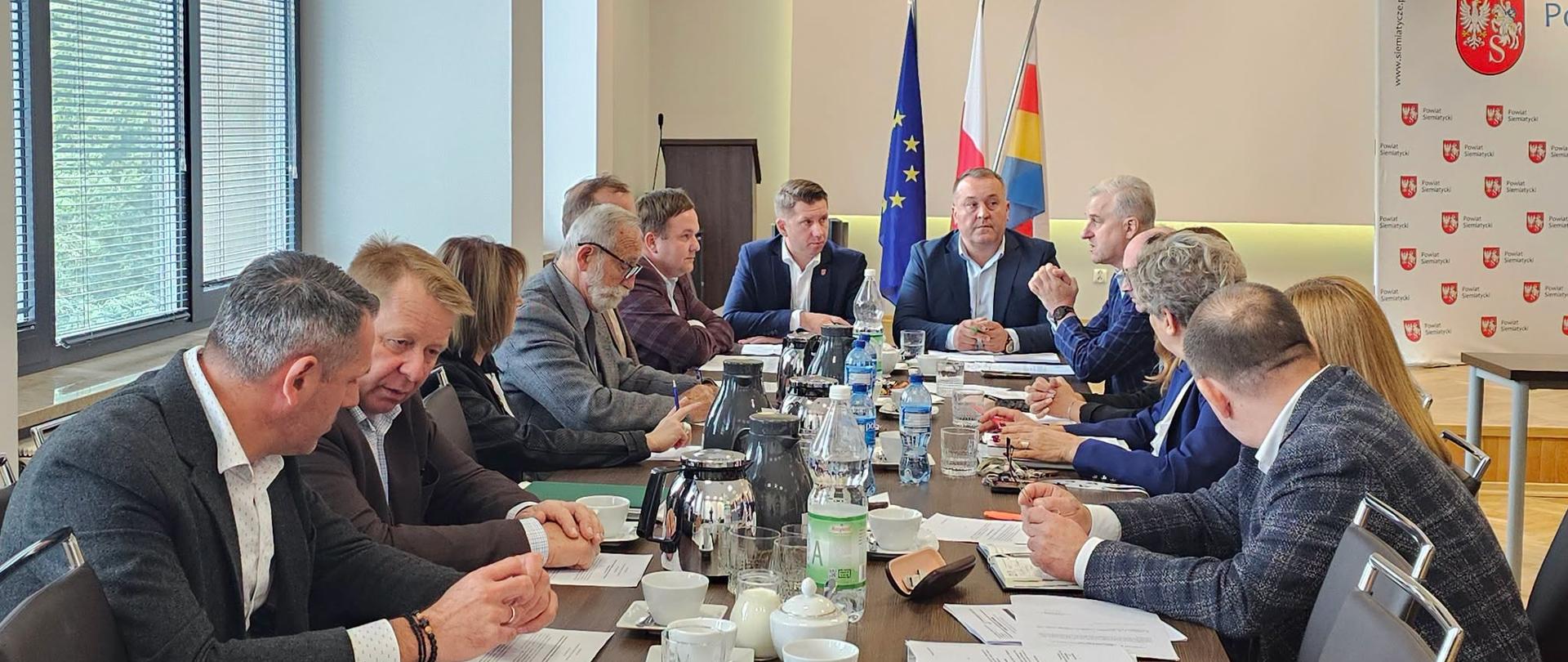 Spotkanie odbywające się przy stole konferencyjnym z udziałem różnych osób, z flagami w tle i dokumentami rozłożonymi na stole.