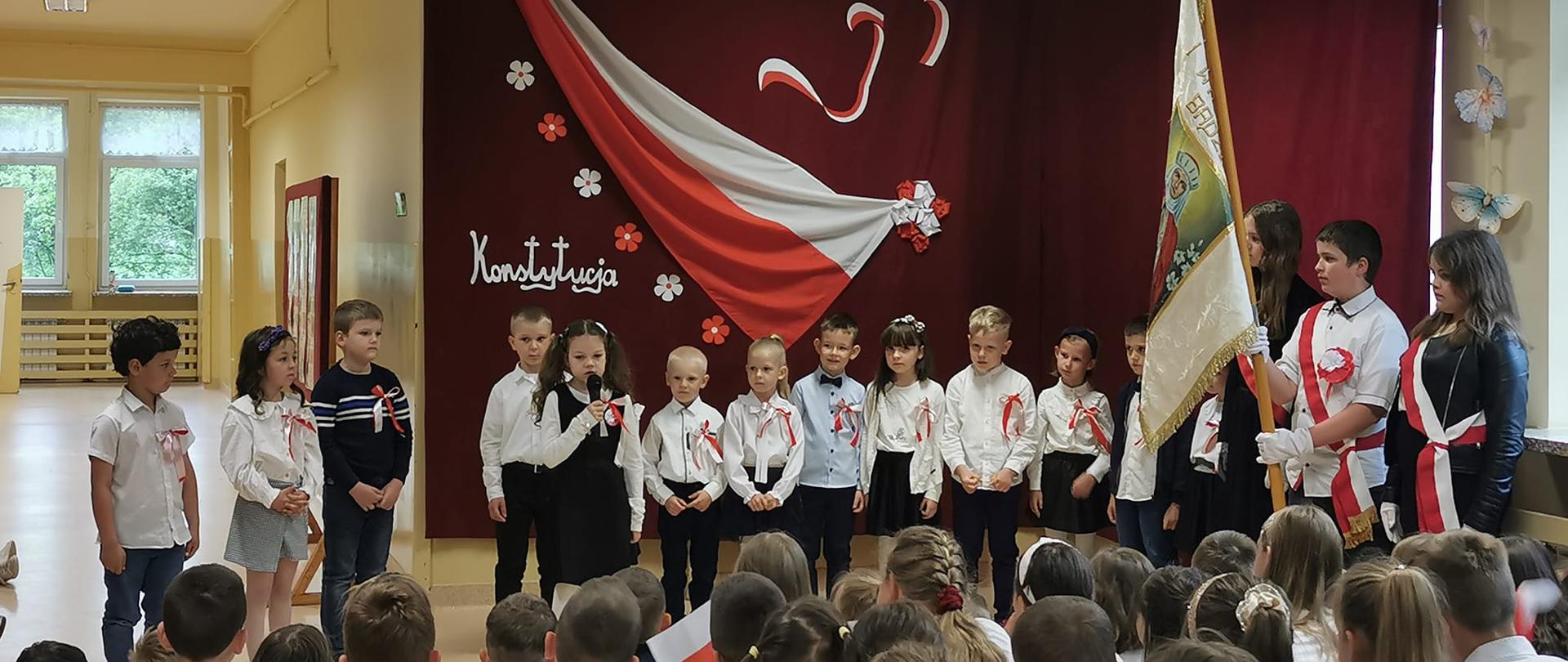 Przedszkolaki ubrani na galowo czyli białe koszule, granatowe spodnie lub spódniczki występują w akademii, na koszulkach mają przypięte biało-czerwone kotyliony. Stoją na tle bordowej zasłony, na której przypięte są biało-czerwone symbole Polski czyli flaga, orzeł w złotej koronie, kwiaty oraz napisy "3 maj" i "Konstytucja". Po prawej stronie stoi poczet sztandarowy ze sztandarem szkoły, przed występującymi widać siedzących uczniów jako widzów