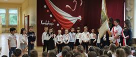 Przedszkolaki ubrani na galowo czyli białe koszule, granatowe spodnie lub spódniczki występują w akademii, na koszulkach mają przypięte biało-czerwone kotyliony. Stoją na tle bordowej zasłony, na której przypięte są biało-czerwone symbole Polski czyli flaga, orzeł w złotej koronie, kwiaty oraz napisy "3 maj" i "Konstytucja". Po prawej stronie stoi poczet sztandarowy ze sztandarem szkoły, przed występującymi widać siedzących uczniów jako widzów