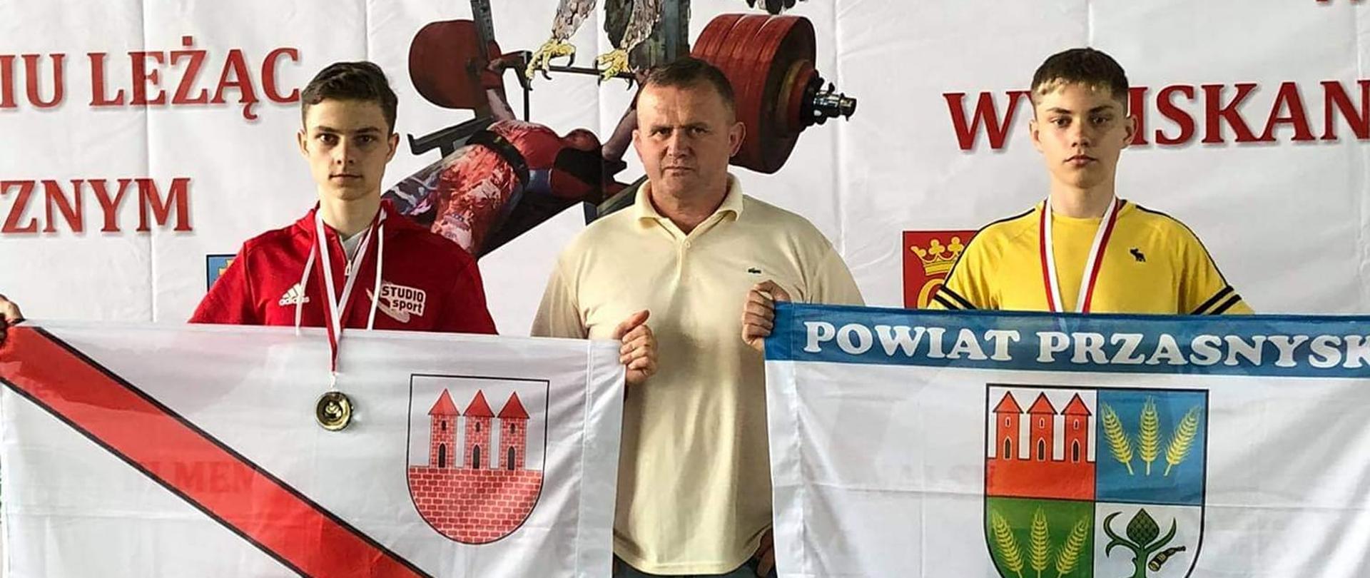 Zdjęcie przedstawia 3 mężczyzn stojących obok siebie, patrząc od prawej strony w czerwonym dresie Hubert Bońkowski trzymający flagę z herbem Miasta Przasnysz, w jasno żółtej koszuli trener Cezary Pykała, w żółtej koszuli Jakub Bońkowski trzymający flagę z herbem Powiatu Przasnyskiego. Na podłodze pokrytej zieloną wykładziną, u stóp zawodników i trenera stoją puchary kolor złotego. 
