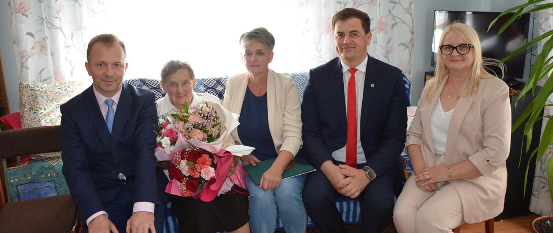 Zdjęcie przedstawia pięć osób pozujących do wspólnej fotografii w przytulnym, domowym wnętrzu. W centrum znajduje się starsza kobieta trzymająca bukiety kwiatów oraz zieloną teczkę. Towarzyszą jej dwaj mężczyźni ubrani w eleganckie garnitury i dwie kobiety w jasnych żakietach. W pomieszczeniu widoczne są kwiatowe zasłony, rośliny doniczkowe, telewizor i stół nakryty obrusem. Uczestnicy spotkania uśmiechają się do obiektywu. Fotografia przedstawia uroczyste, serdeczne spotkanie w kameralnej atmosferze.