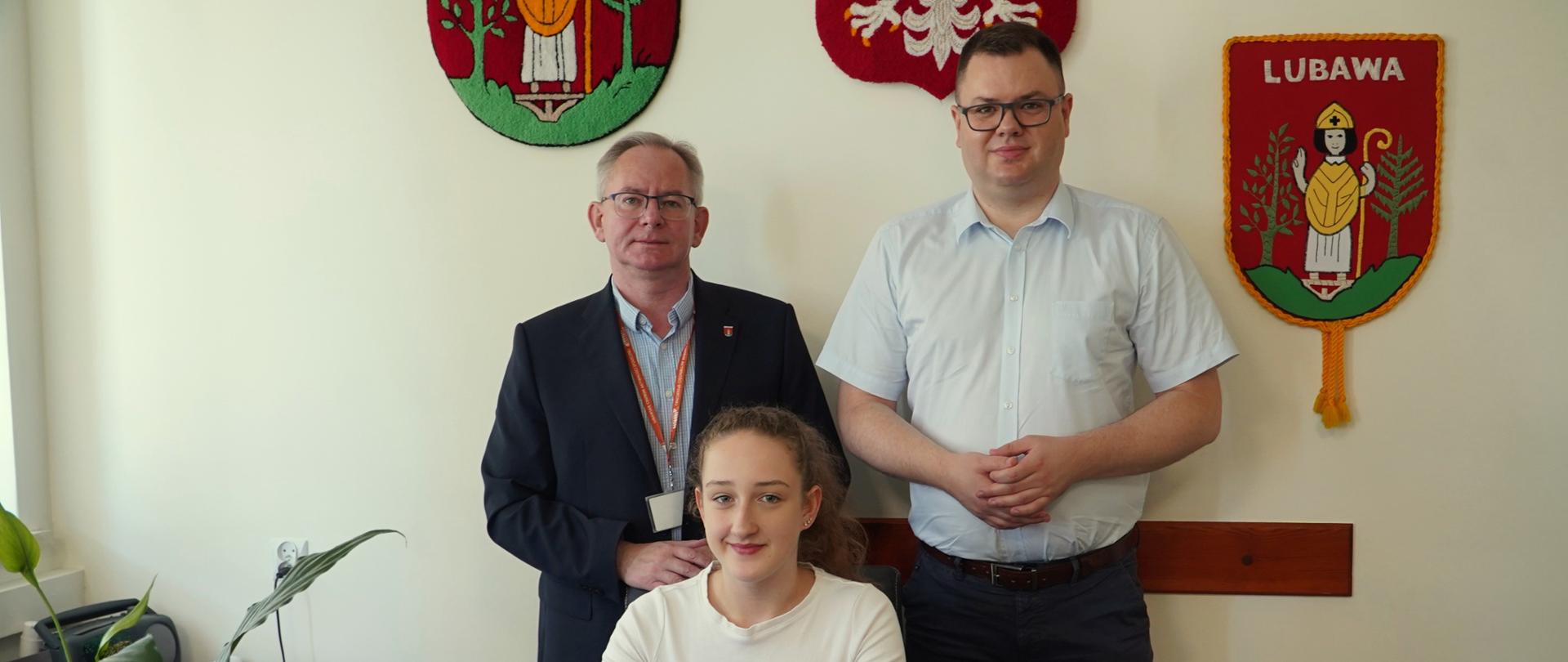 Dziewczynka siedzi przy biurku, a za nią dwaj mężczyźni, jeden w ciemnym garniturze, drugi w jasnej koszuli.