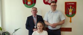 Dziewczynka siedzi przy biurku, a za nią dwaj mężczyźni, jeden w ciemnym garniturze, drugi w jasnej koszuli.