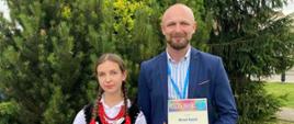 dziewczyna w ludowym kolorowym stroju trzyma w ręku dyplomy obok niej mężczyzna w dżinsach i ciemnej marynarce 