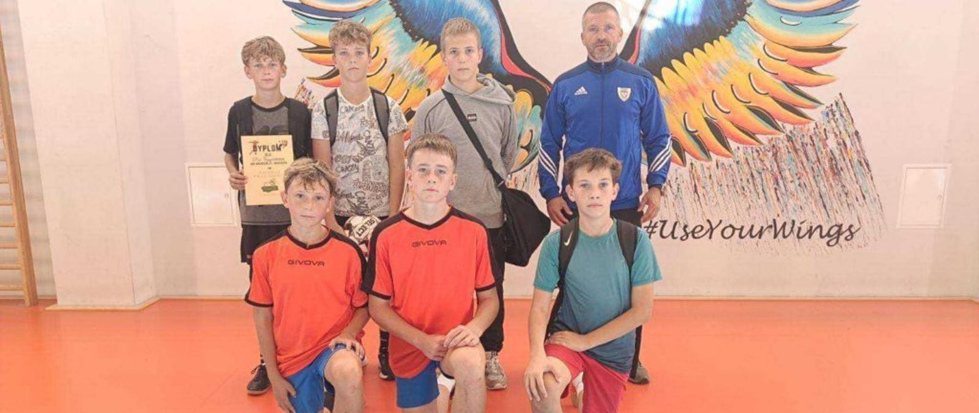 Trzech chłopców przyklęka na jednym kolenie. Za nimi stoi trener z trzema zawodnikami. Wszystko na tle skrzydeł namalowanych na ścianie hali sportowej w Łapczycy gdzie odbywały się zawody.