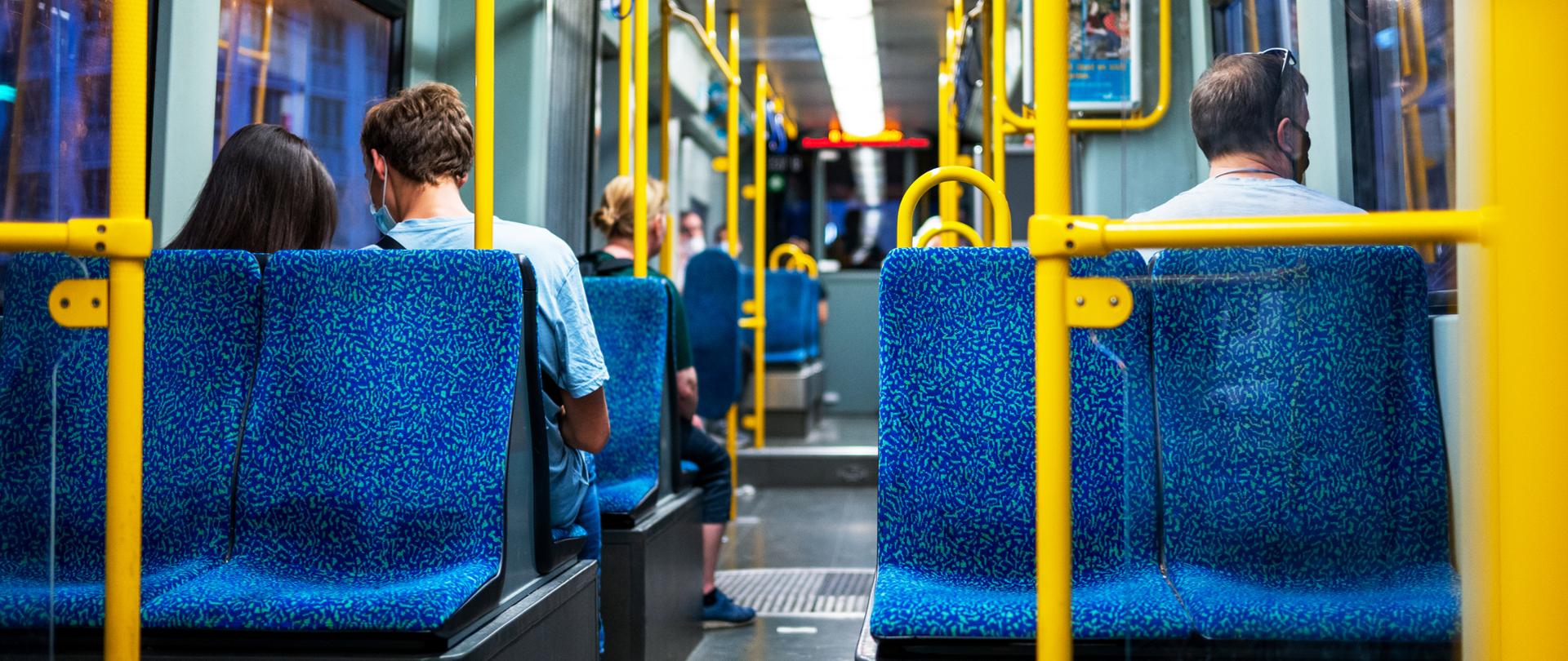 Widok wnętrza autobusu, niebieskie fotel, żółte poręcze. Na siedzenia cztery osoby spoglądające w okno, plecami do widza.