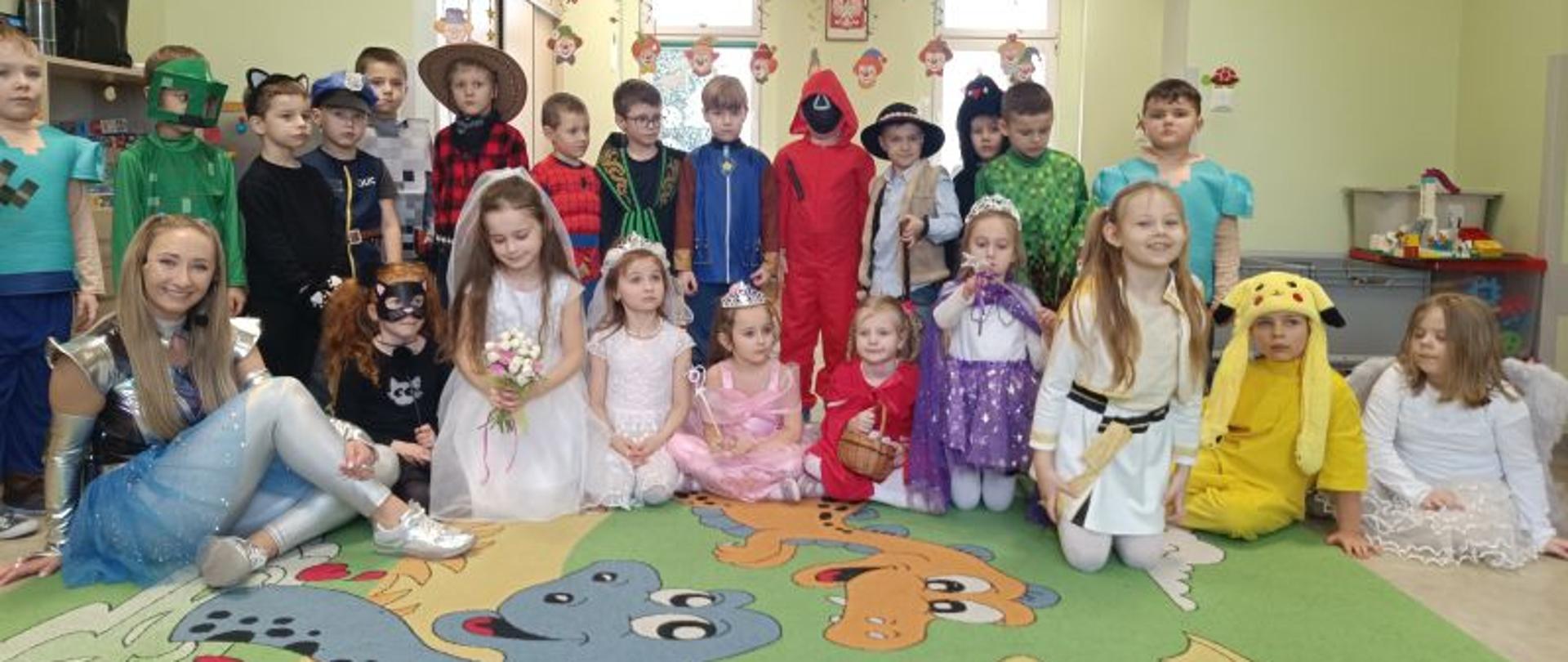 Dzieci z grupy Misie prezentują swoje stroje karnawałowe. Obok siedzi animatorka. W tle widać dekoracje sali i dwa okna. 