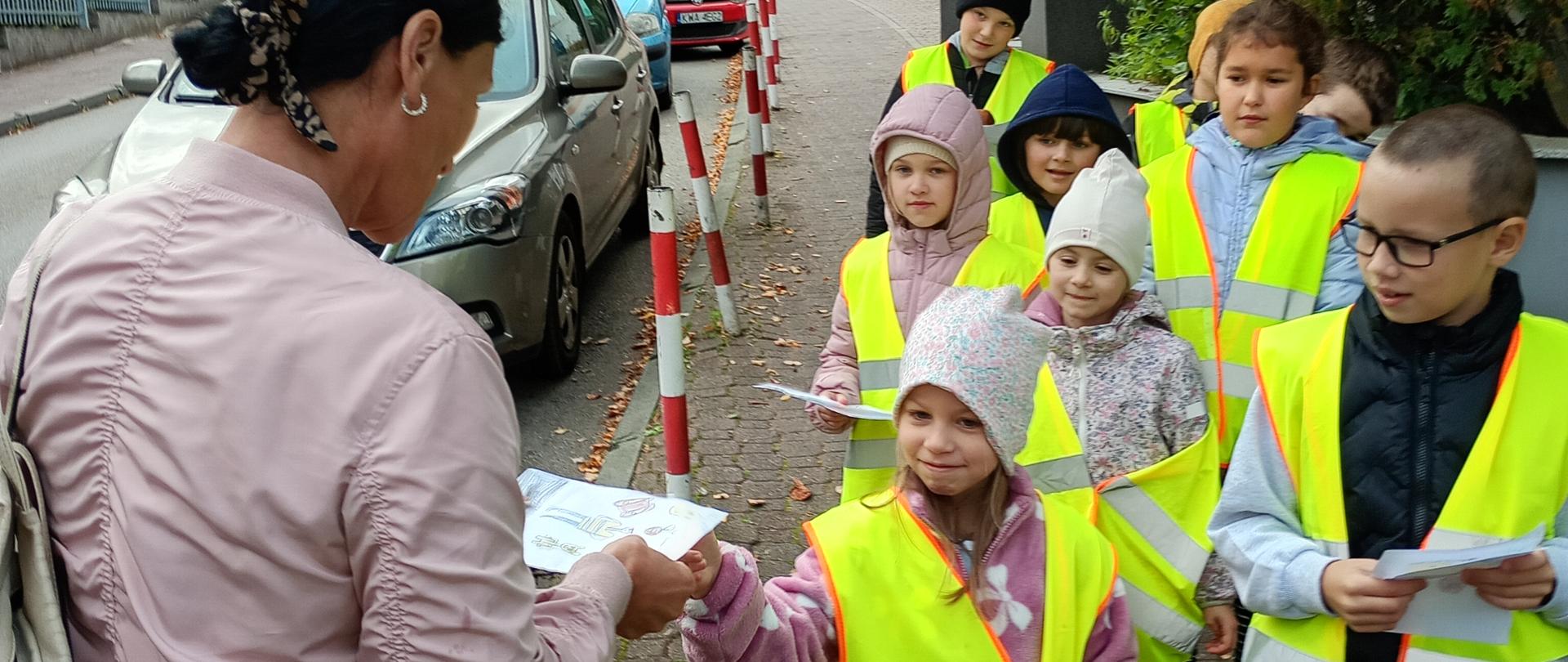 Na chodniku przy ulicy stoi grupa dzieci w jaskrawych kamizelkach odblaskowych. Dziewczynka z przodu wręcza kartkę kobiecie stojącej tyłem do aparatu, a pozostałe dzieci trzymają w rękach kartki i czekają obok. W tle widać zaparkowane samochody, słupki przy chodniku, drzewa oraz budynki.