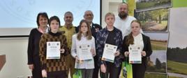 Zdjęcie grupowe laureatów z grupy IV - szkoły ponadpodstawowe. Na fotografii młodzież, która w rękach trzyma dyplomy oraz torby z nagrodami. Wraz z nimi na zdjęciu starosta sokołowski, przewodniczący Rady Powiatu Sokołowskiego, prezes oddziału powiatowego ZOSP RP i zastępca komendanta KP PSP w Sokołowie Podlaskim.