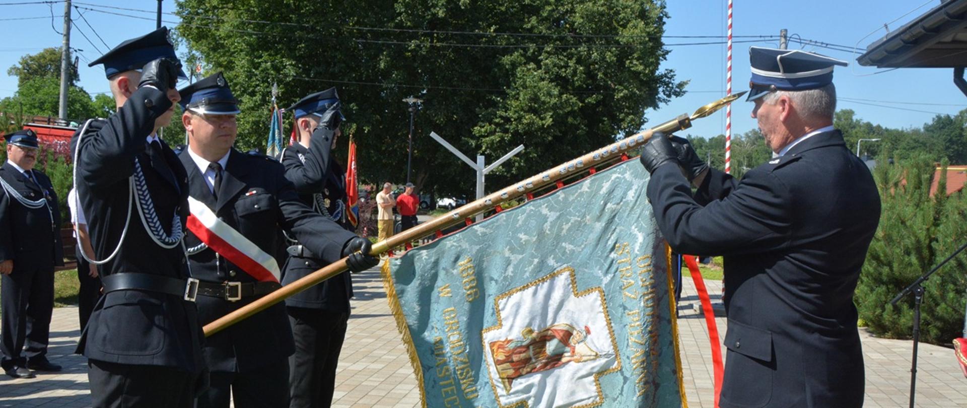 trzech strażaków w galowych strojach stoją przy sztandarze czwarta zakłada na sztandar odznaczenie 