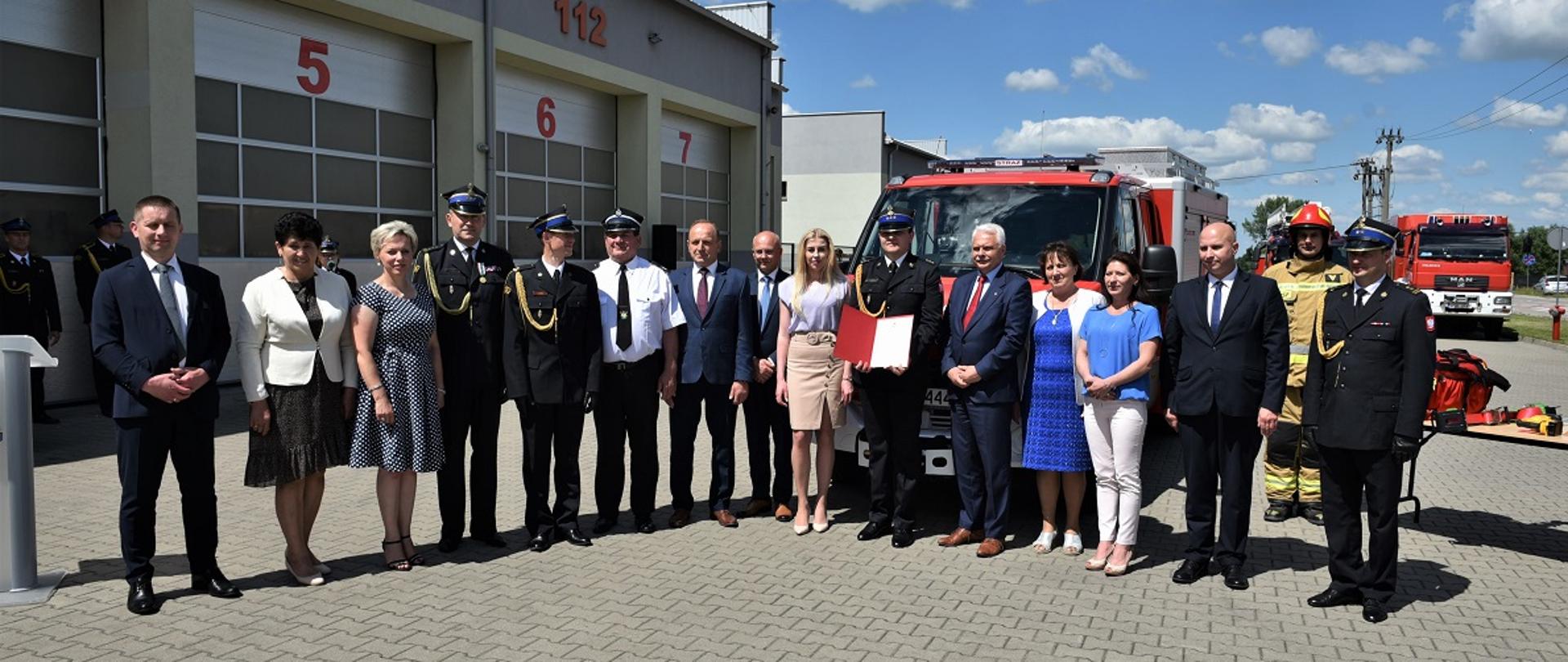 Zdjęcie grupowe samorządowców na tle garaży i nowego lekkiego samochodu ratownictwa technicznego.