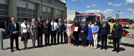 Zdjęcie grupowe samorządowców na tle garaży i nowego lekkiego samochodu ratownictwa technicznego.