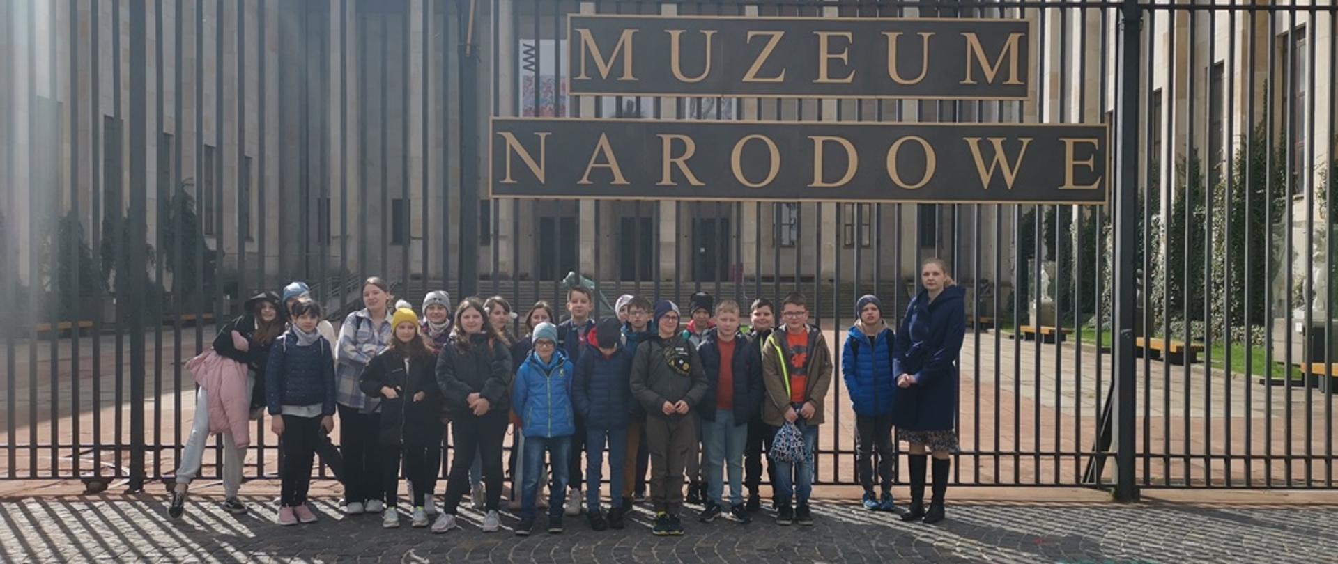 Grupa dzieci stojąca przed bardzo wysokim ogrodzeniem z napisem Muzeum narodowe