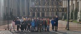 Grupa dzieci stojąca przed bardzo wysokim ogrodzeniem z napisem Muzeum narodowe
