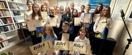 Pamiątkowe zdjęcie grupowe uczestników, laureatów oraz organizatorów konkursu recytatorskiego w bibliotece. Dzieci i młodzież trzymają w rękach dyplomy, torby z upominkami oraz symboliczne duże „Bilety” za zajęcie czołowych miejsc.