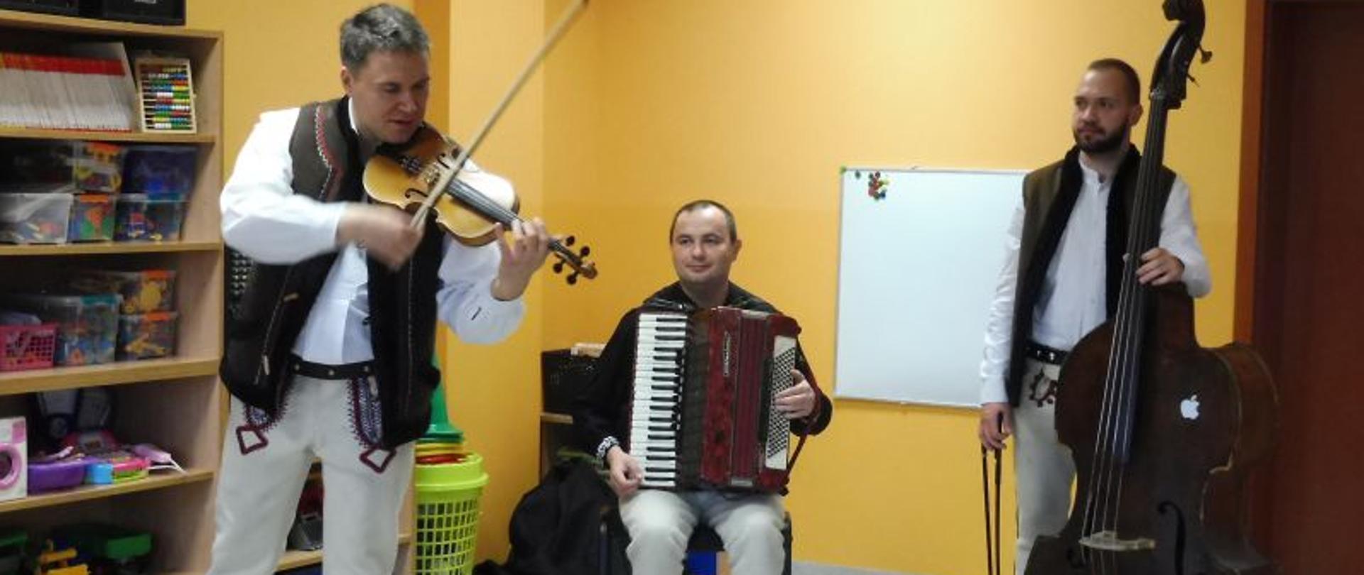 Trzech mężczyzn w strojach góralskich z instrumentami: skrzypce, akordeon, kontrabas. 