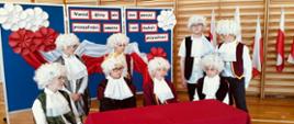 Chłopcy przebrani za magnatów - twórców konstytucji w perukach na głowie, siedzą przy stole i obradują, za nimi na ściance dekoracyjnej na granatowym tle napis "narów, który nie zna swojej przeszłości, umiera i nie buduje przyszłości" oraz biało-czerwone kwiaty