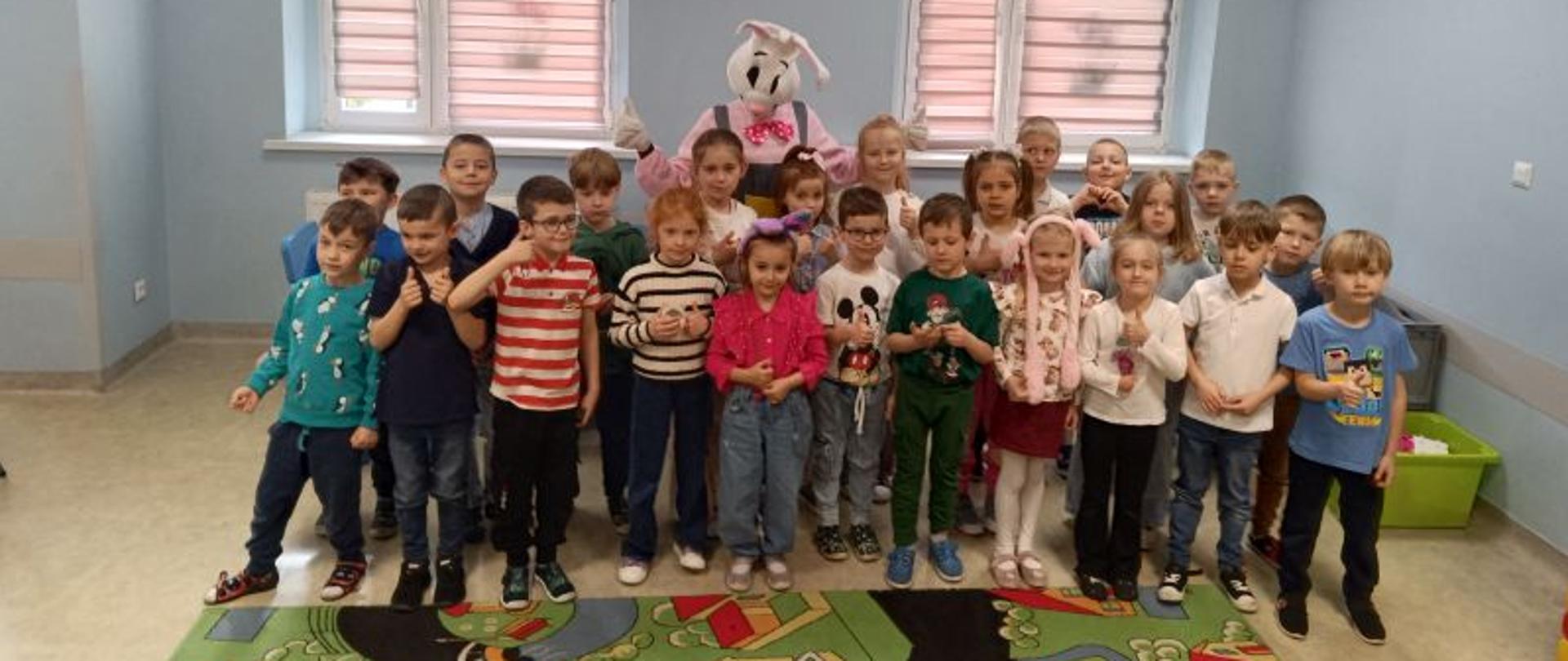 Dzieci w wieku przedszkolnym stoją w dwóch szeregach, za nimi postać zajączka. W tle widać dwa okna zasłonięte roletami, miedzy nimi wisi godło Polski. Na podłodze leży kolorowy dywan. 