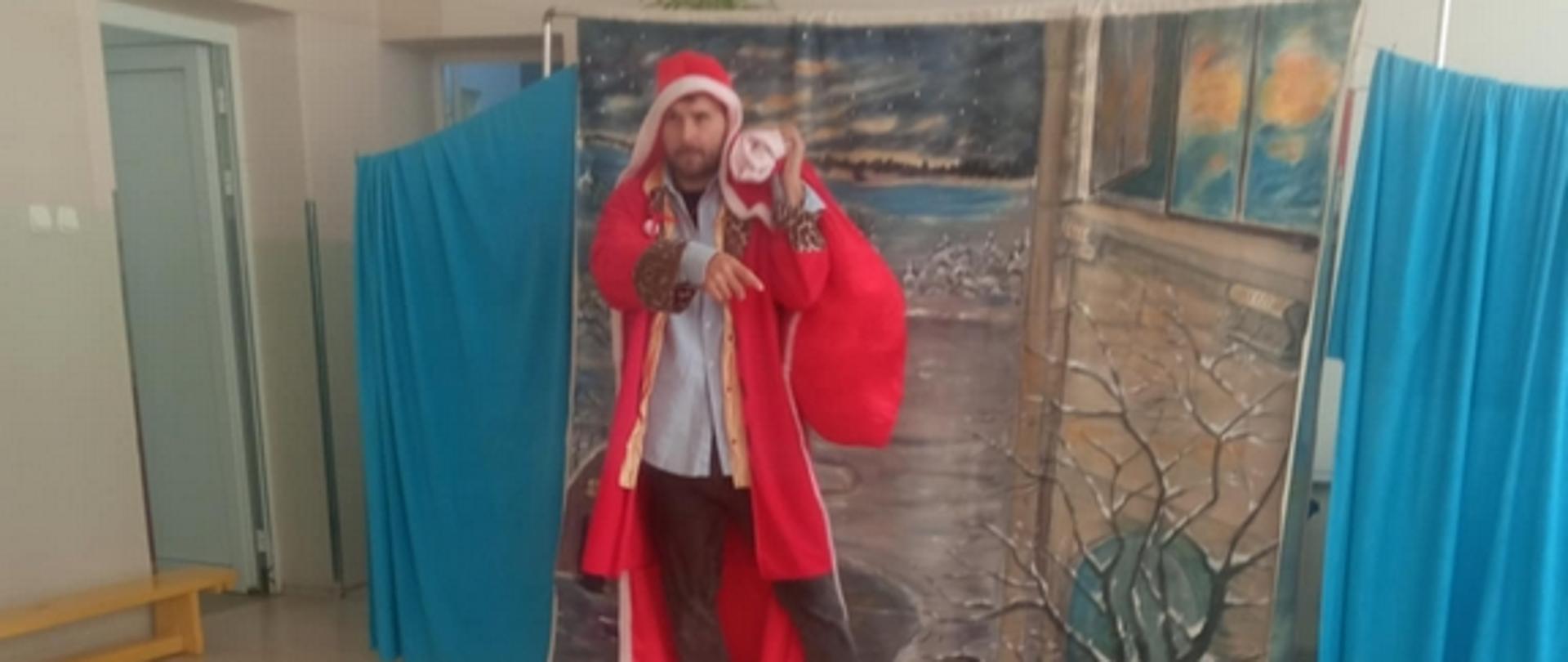 W sali przedszkolnej rozłożona dekoracja. Przed nią aktor w przebraniu Mikołaja trzymając wielki czerwony wór na lewym ramieniu. W pierwszym kadrze widać głowy siedzących dzieci zwrócone w kierunku aktora.