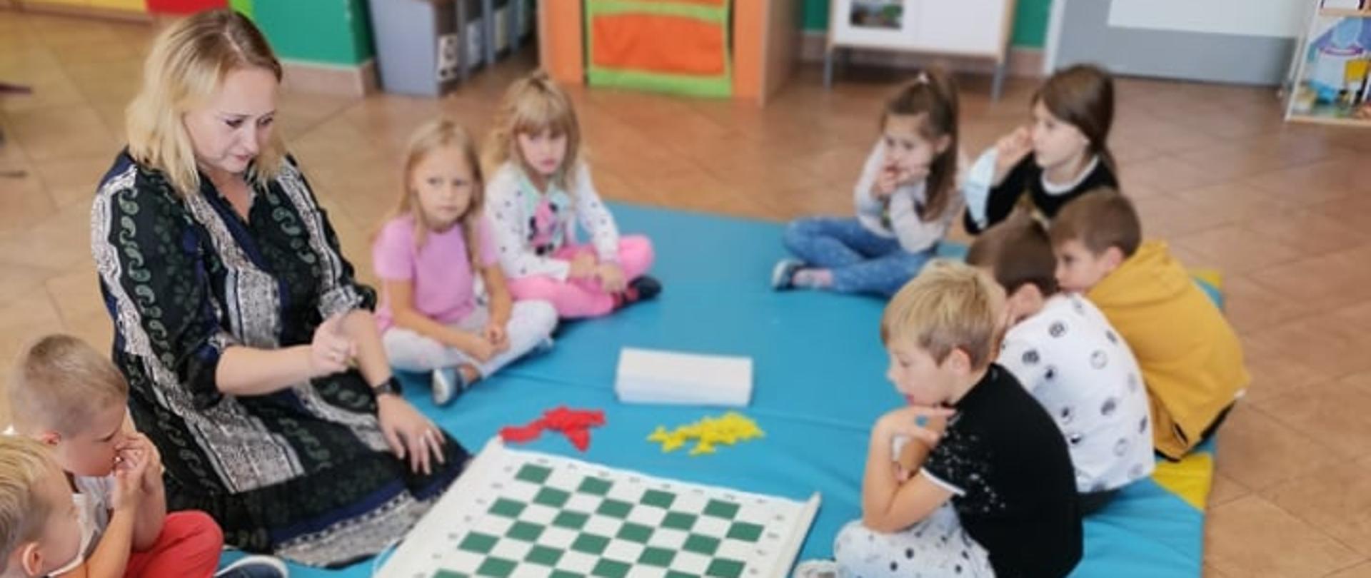 Dzieci siedzą w kole na niebieskim materacu. Na środku leży szachownica.
