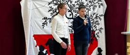 Uczeń przemawia do mikrofonu, obok stroi uczennica, w tle dekoracja: na białym płótnie biało-czerwona mapa polski, na niej czarna sylweta drzewa z huśtającą się na huśtawce dziewczynką