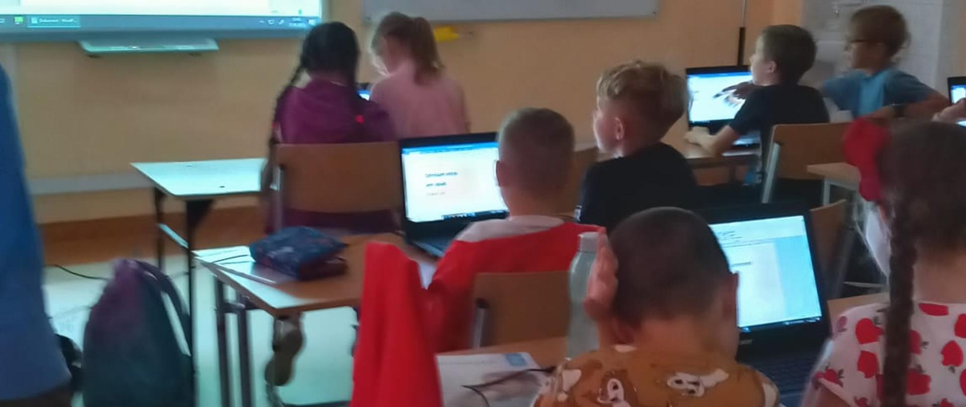 Sala komputerowa, na górze po lewej stronie tablica multimedialna, po prawej stronie biała tablica. Poniżej uczniowie siedzący w ławkach, patrzący w ekrany laptopów. 