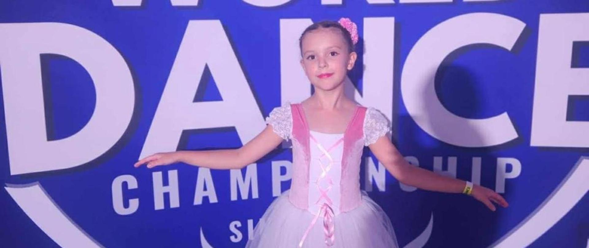 Antosia stoi w sukni baletowej, za nią na niebieskim tle napis "World Dance Championship", nad nim 5 gwiazdek, środkowa największa