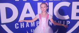 Antosia stoi w sukni baletowej, za nią na niebieskim tle napis "World Dance Championship", nad nim 5 gwiazdek, środkowa największa