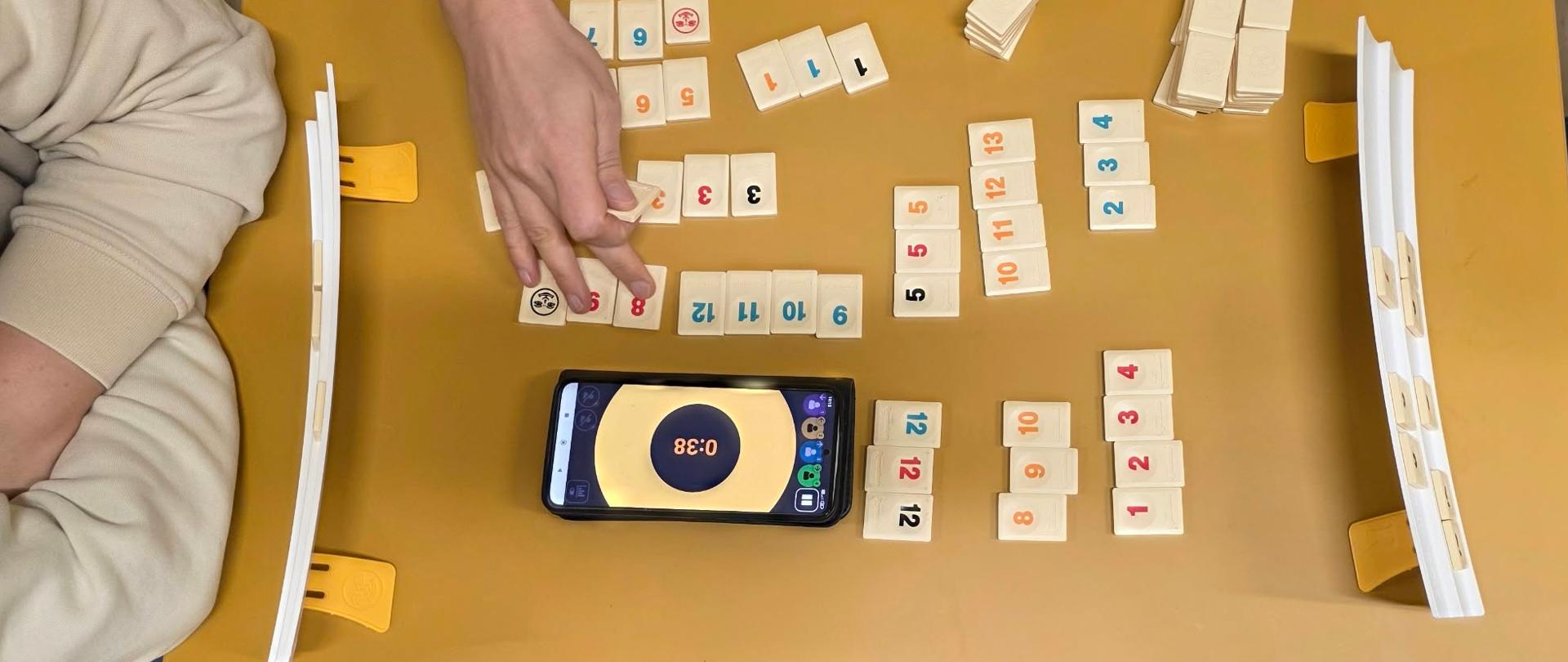 Przykładowa rozgrywka w Rummikub: na stole stanowiska graczy z płytkami oraz aplikacja na telefonie komórkowym