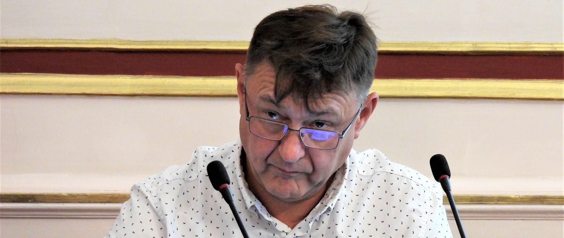 Gerard Nickel - mężczyzna w średnim wieku spogląda spod okularów. Siedzi przy stole w czasie sesji Rady Gminy Świerklaniec, która odbywa się w jednej z sal w Pałacu Kawalera. Przed mężczyzną ustawione są mikrofony i szklane butelki z wodą.