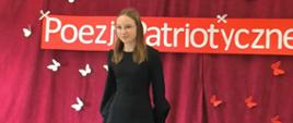 Uczennica odświętnie ubrana recytuje poezję patriotyczną, w tle na bordowej kotarze biały napis "Konkurs Recytatorski Poezji Patriotycznej" oraz przypięte papierowe białe motylki