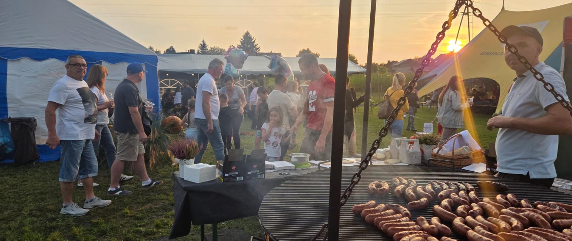 Mężczyzna przy grillu pełnym kiełbasek w tle ludzie i namioty na łące