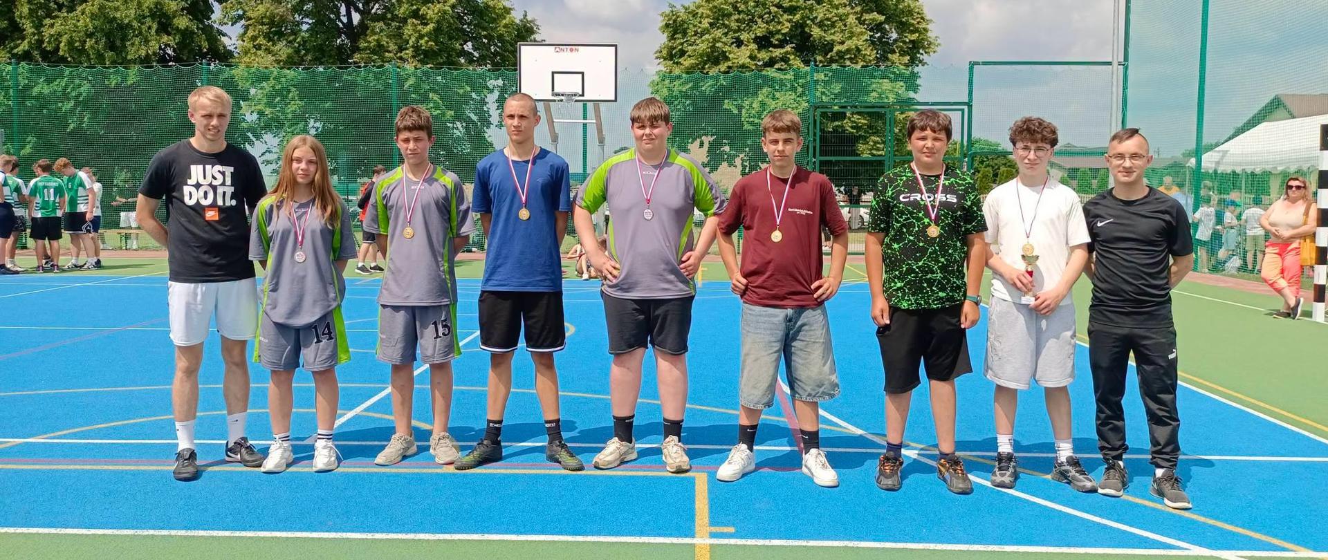 Dziewięcioro nastolatków (jedna dziewczyna i ośmiu chłopców) stoi w rzędzie na niebiesko-zielonym boisku sportowym. Większość z nich ma na szyi medale, a jeden trzyma mały puchar. W tle widać siatkę ogrodzeniową, kosz do koszykówki oraz drzewa pod zachmurzonym niebem.