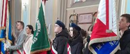 W budynku kościoła stoi troje uczniów naszej szkoły (chłopak i dwie dziewczyny.), reprezentujący sztandar szkoły. Uczniowie ubrani są w stroje galowe, na ramionach mają przewieszone biało-czerwone szarfy.