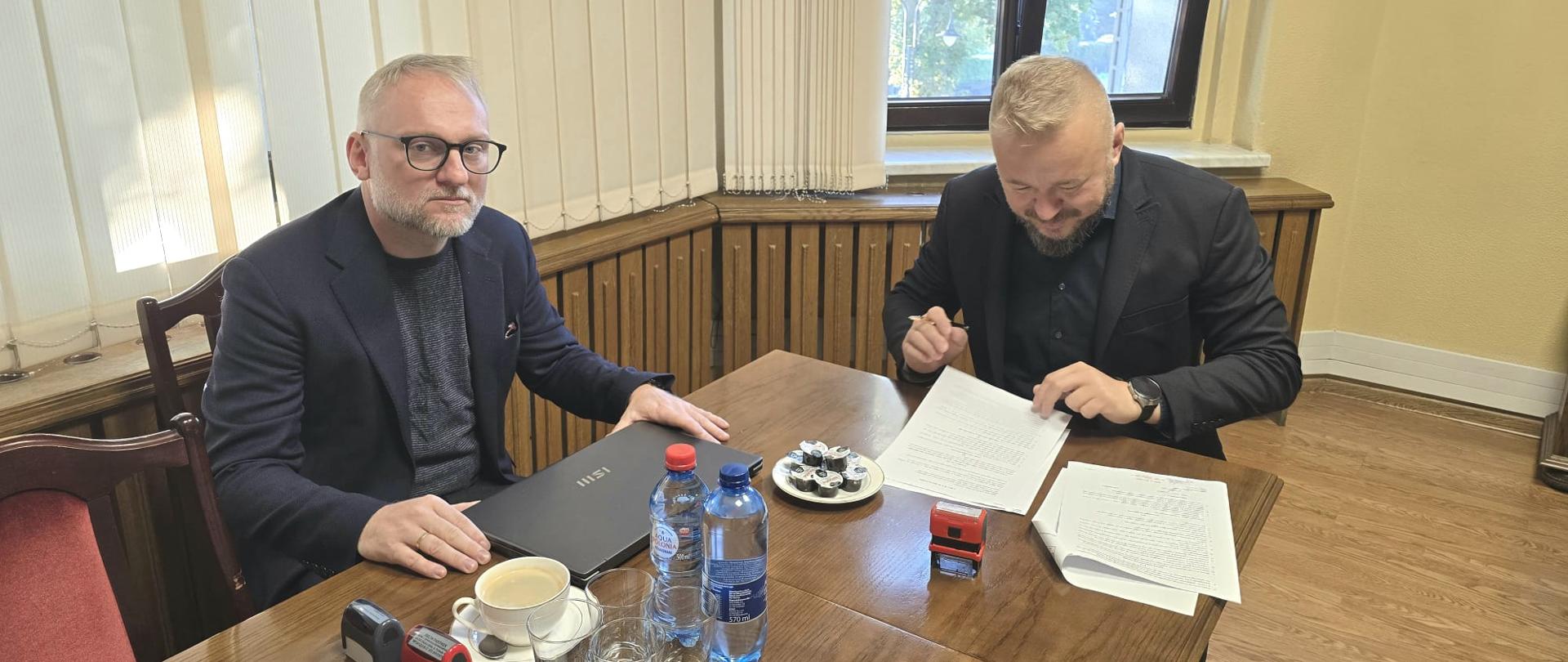 Dwóch mężczyzn siedzi przy stole, jeden podpisuje dokumenty.